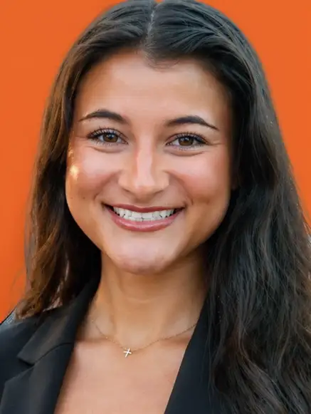 Young man with curly dark hair wearing blue scrubs and white undershirt smiling against an orange background.